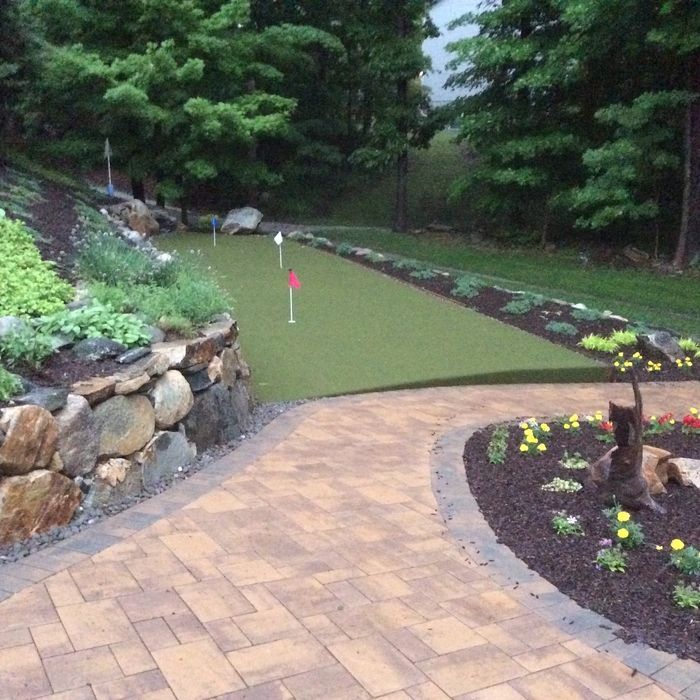 A backyard putting green with several flags, adjacent to a stone retaining wall and a paved walkway with flower beds.