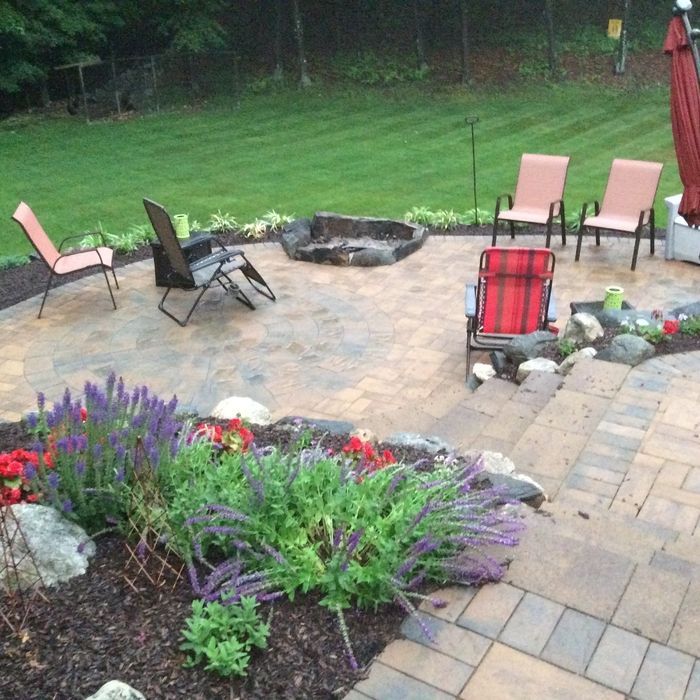 A stone patio with a fire pit, several outdoor chairs, and garden beds with purple flowers in the foreground.