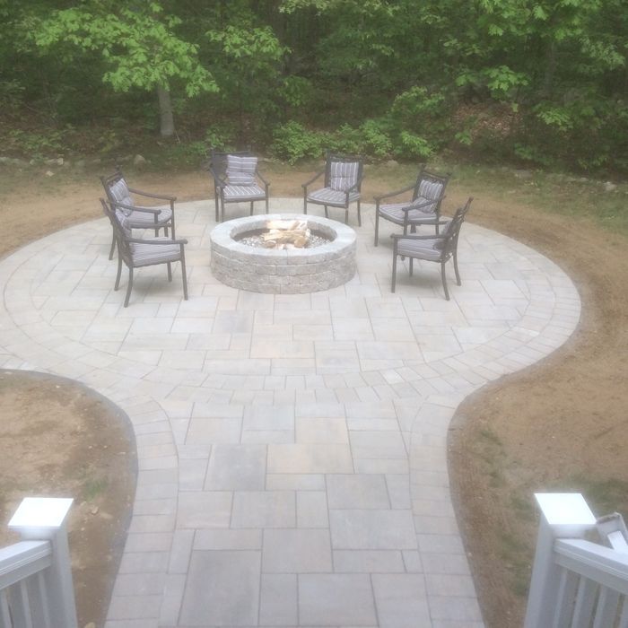 A circular stone patio features a central fire pit surrounded by six gray chairs, set against a backdrop of green trees.