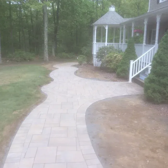 A curved stone paver walkway leads to a porch with a gazebo-style roof, set against a background of trees.