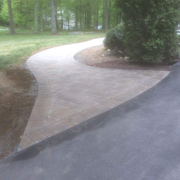 A curved paver walkway transitions from a dark asphalt driveway through a grassy, wooded yard.