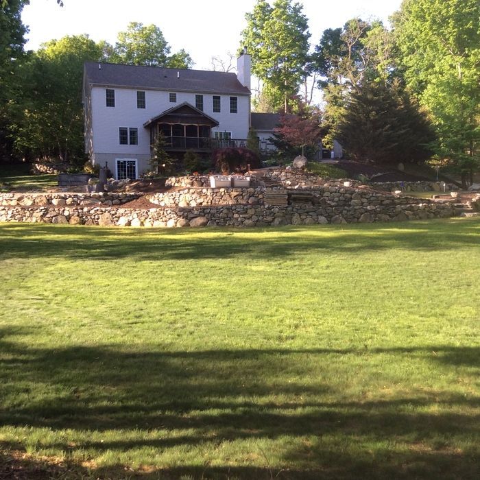 A two-story white house with a screened porch sits behind a tiered stone retaining wall overlooking a large green lawn.
