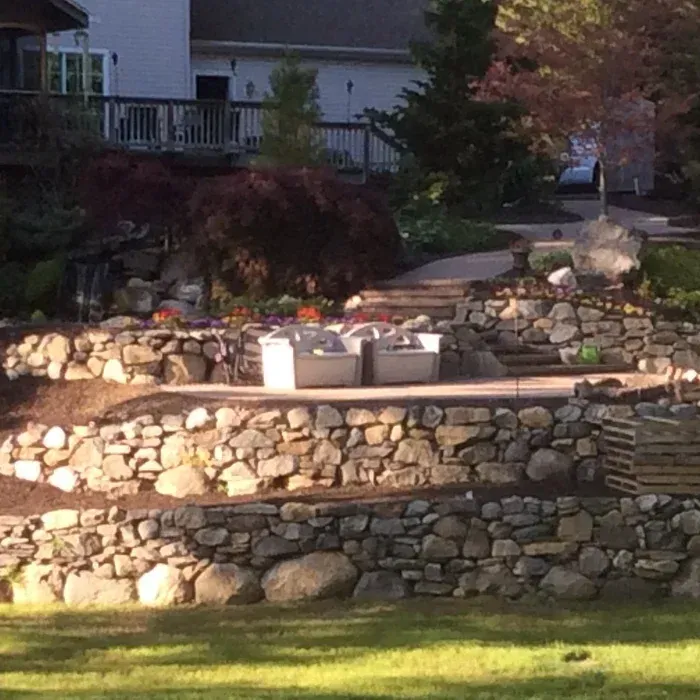 A multi-level backyard landscape featuring stone retaining walls, a patio with chairs, and a paved path leading to a house.