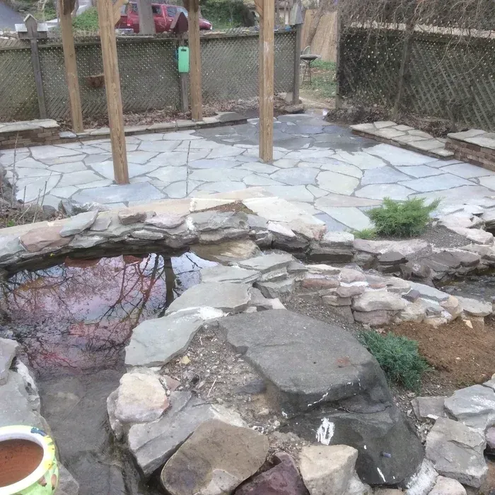 A stone-paved patio with a wooden pergola frame, set behind a small, rock-lined pond in a backyard garden.