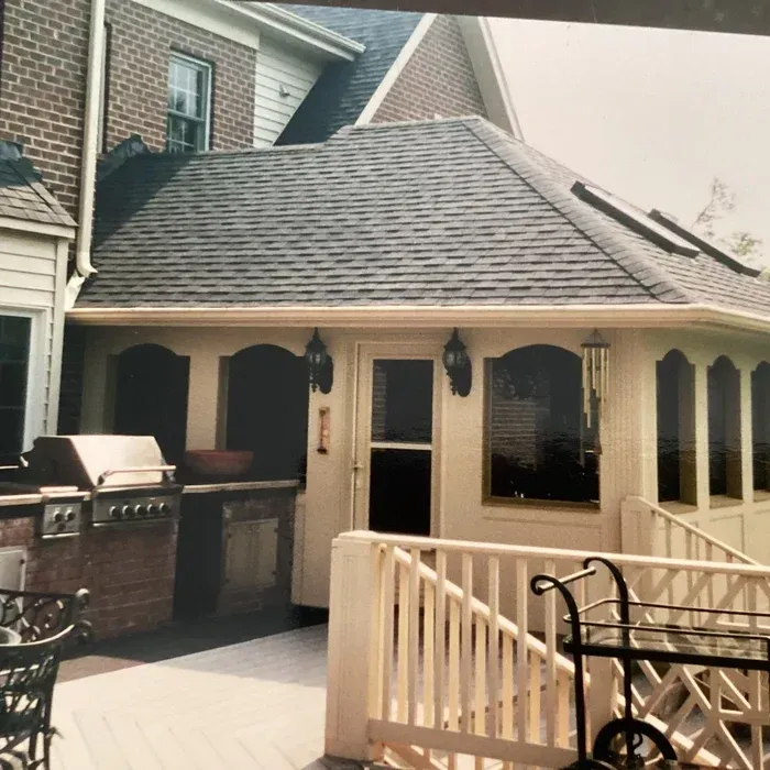 A tan sunroom with a brick outdoor grill, a white railing, and a gray shingled roof attached to a brick house.