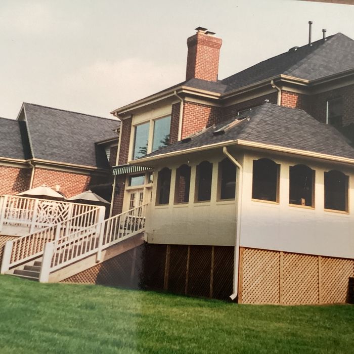 A two-story red brick house with a cream-colored sunroom, white wooden stairs leading to a deck, and a green lawn.