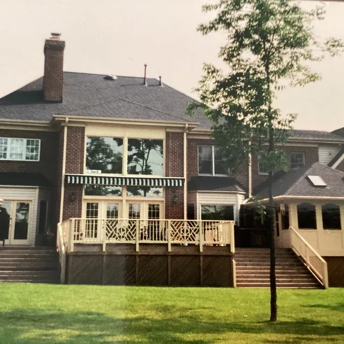 A two-story brick house with a raised wooden deck, large windows, a striped awning, and a sunroom, viewed from a lawn.