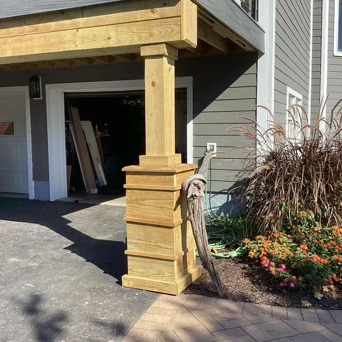 A wooden support post stands on a driveway next to a tan house with a garage, with a gnarled piece of driftwood nearby.