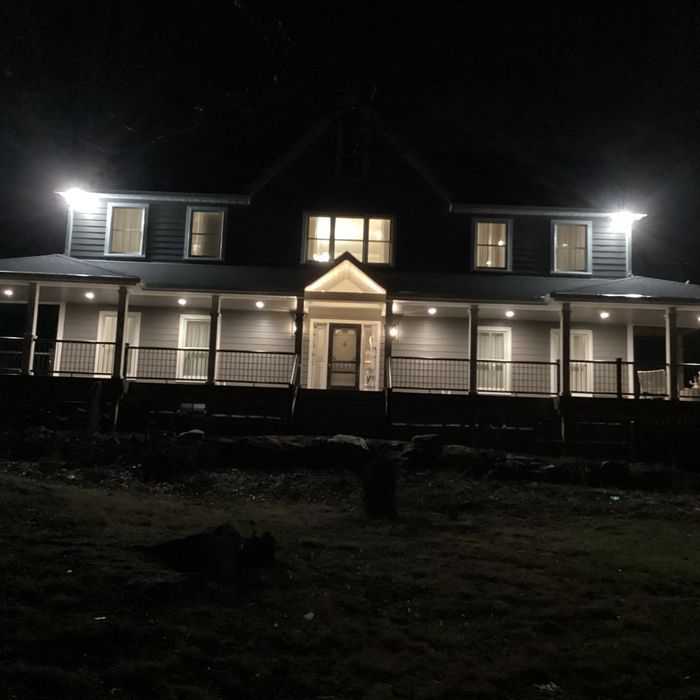 A two-story dark-colored house at night, illuminated by bright exterior porch lights and glowing interior windows.