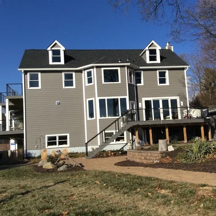 A large gray three-story house with a wooden deck, outdoor stairs, and a backyard landscape under a clear blue sky.