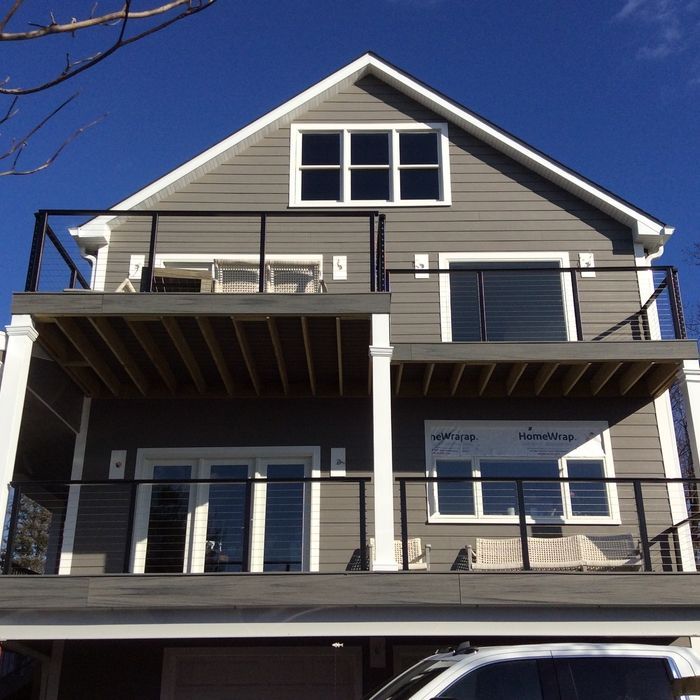 A multi-story gray house with white trim, two balconies, a large deck, and a white vehicle parked in the foreground.