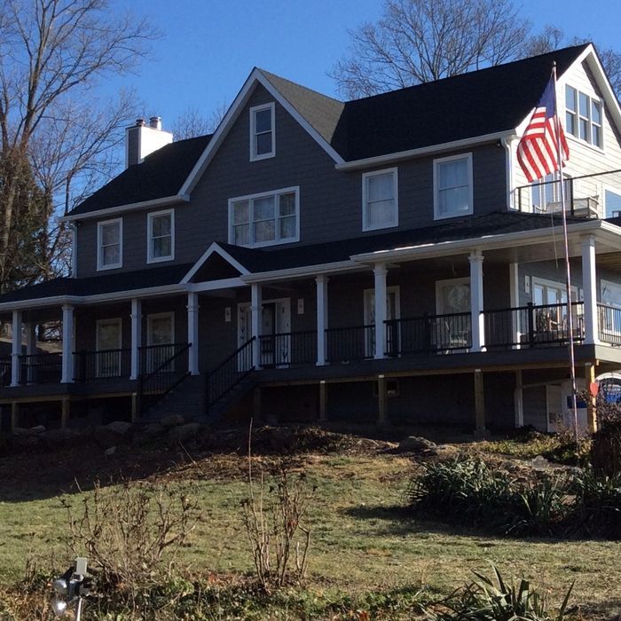 A gray two-story house with a wraparound porch, black railings, a front gable, and an American flag on a flagpole.