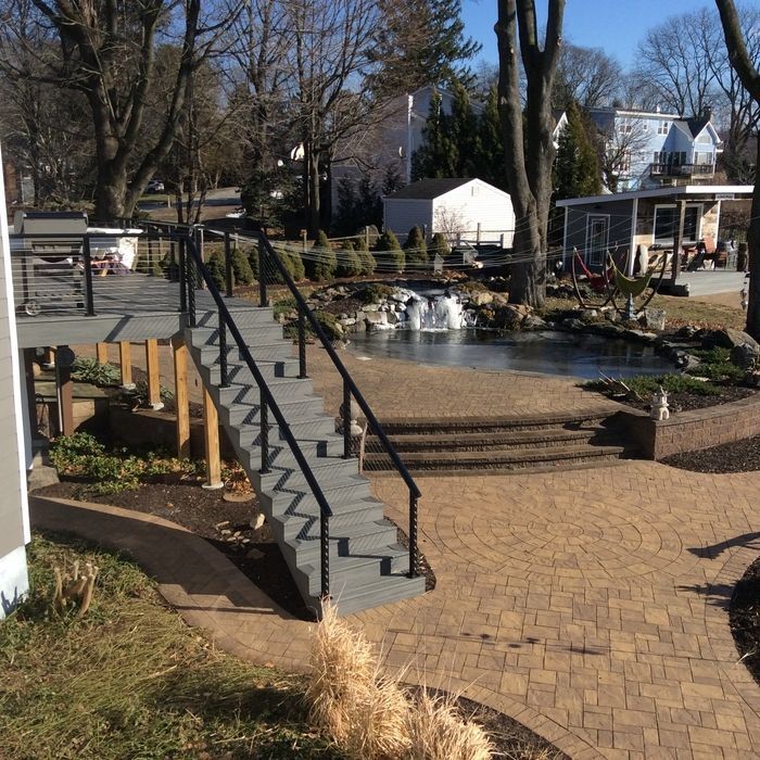 A grey deck with stairs leading down to a stone patio, a small pond with a waterfall, and a backyard in a residential area.