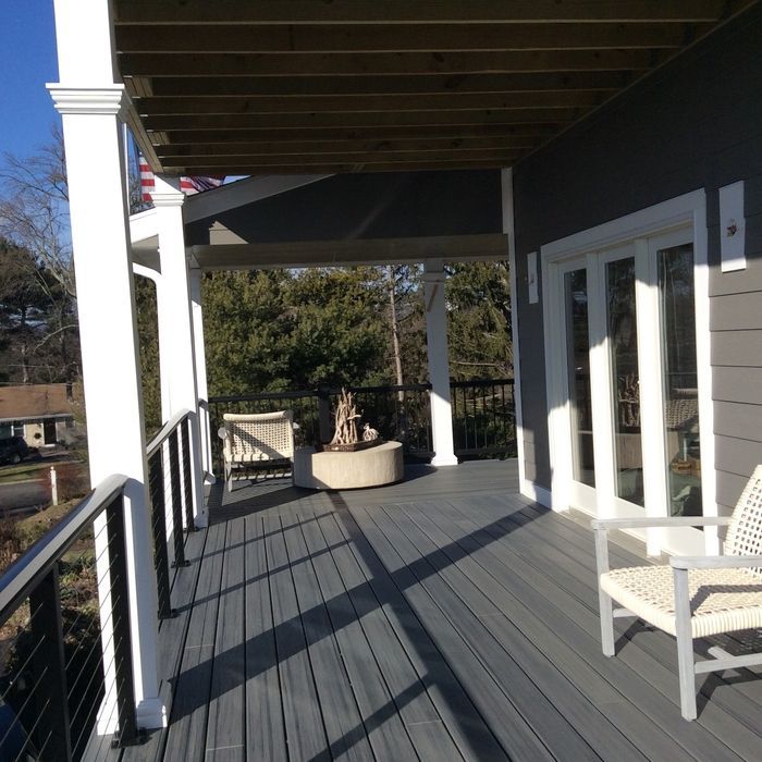 A gray wooden deck with white columns, a cable railing, and chairs under a roof, overlooking a sunny backyard scene.