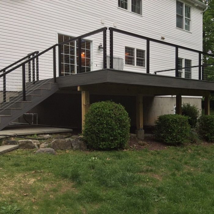 A gray deck with black metal railings and stairs attached to the back of a white house with bushes underneath.