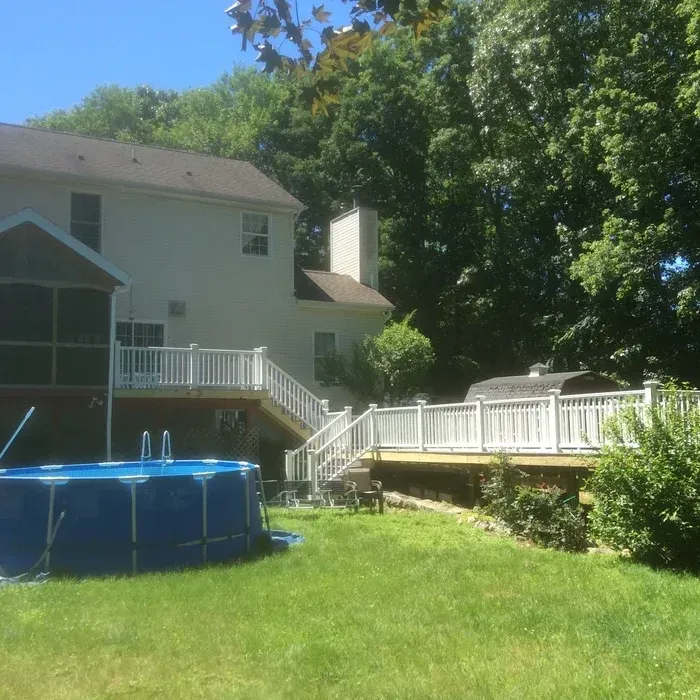 A large backyard with a round blue above-ground swimming pool, a wooden deck, and a multi-story house on a sunny day.
