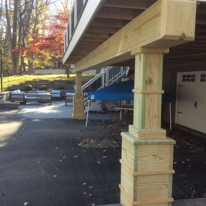 A close-up of a wooden support beam under a deck, with a backyard patio and trees visible in the background.