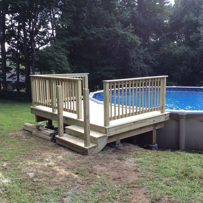 A wooden deck with railings stands beside a blue above-ground swimming pool in a grassy backyard with trees.
