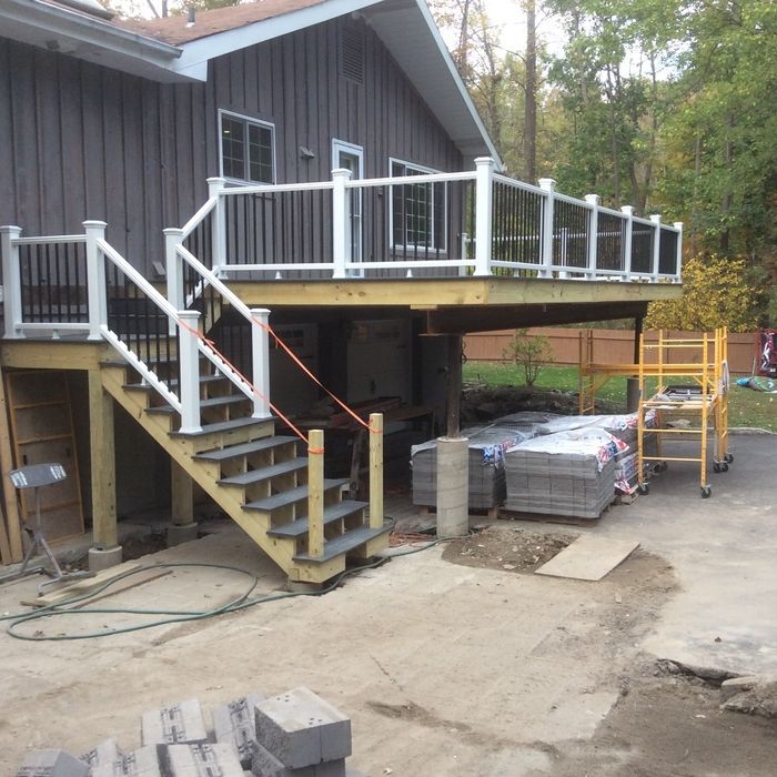 A gray house with a partially constructed elevated wooden deck and stairs featuring white railings and black balusters.