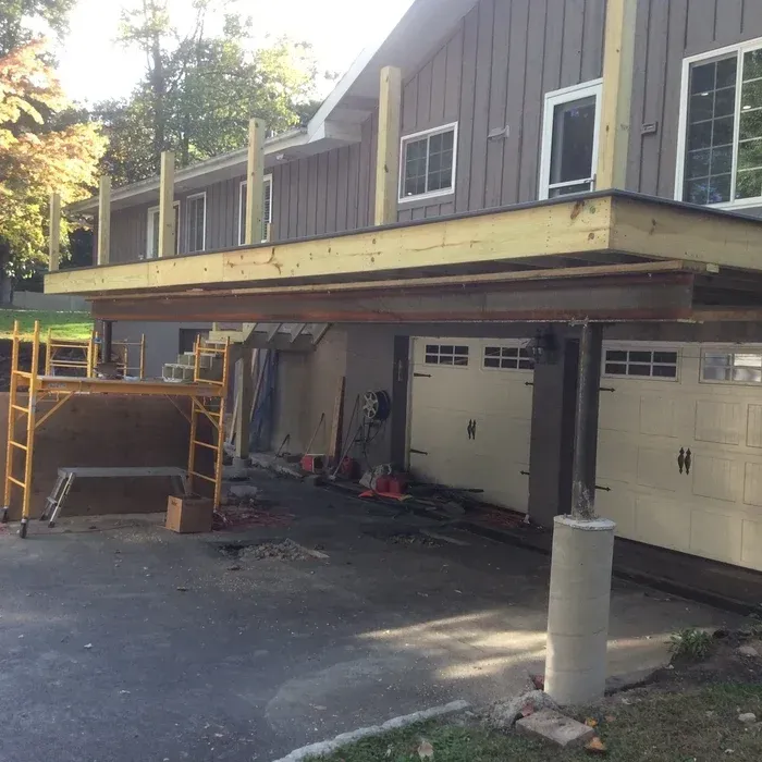 Construction work on a deck attached to the second story of a gray house above a two-car garage driveway.