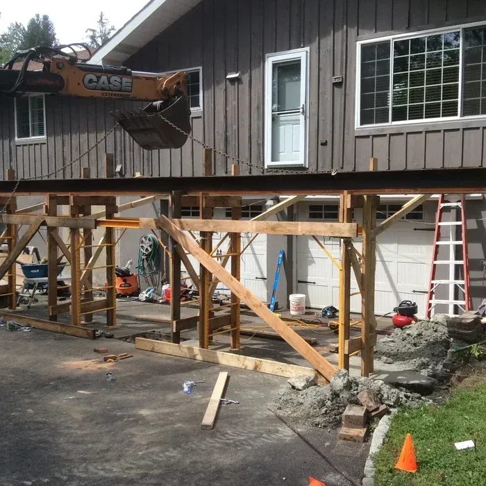 A construction site showing a temporary wooden support structure built in front of a house, with a yellow excavator above.