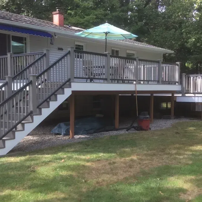 A raised wooden deck with grey railings and stairs stands against a white house, with a light blue umbrella on top.