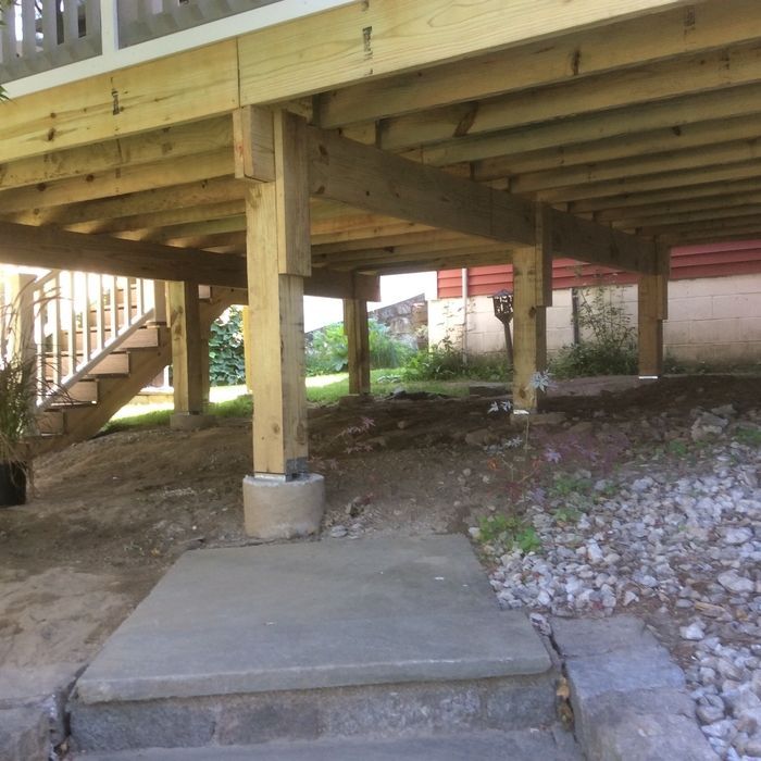 The underside of a raised wooden deck showing support posts resting on concrete footings above a stone patio.