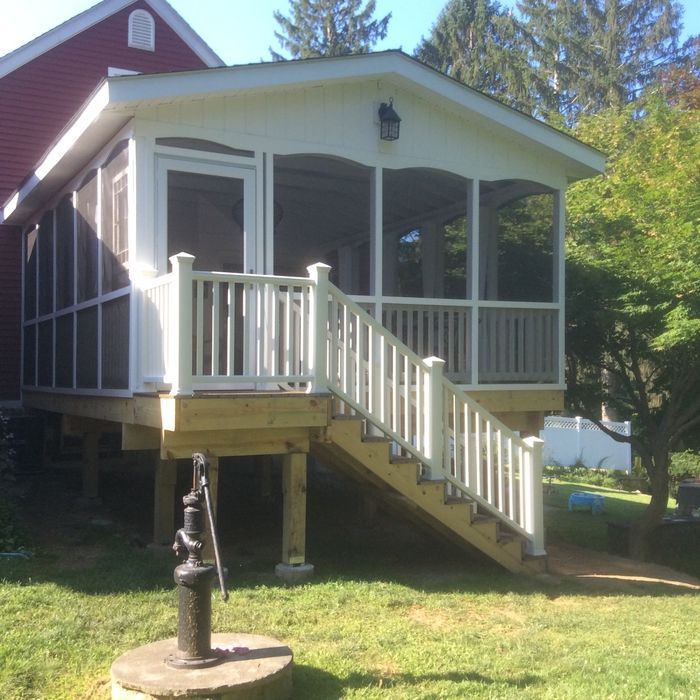 A white screened-in porch with a staircase attached to a red house, positioned above a grass lawn with an old water pump.