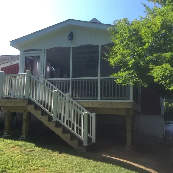 A wooden deck with white railings and stairs leading up to an elevated, white-framed screened porch on a sunny day.