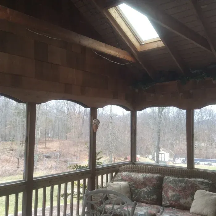 A view from inside a covered porch with wooden walls, a skylight, and outdoor seating looking out onto a wooded area.