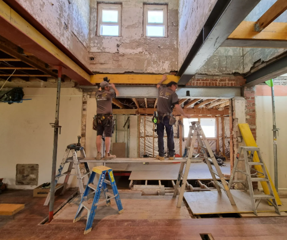 Two workers on ladders framing a renovation in a tall, unfinished room