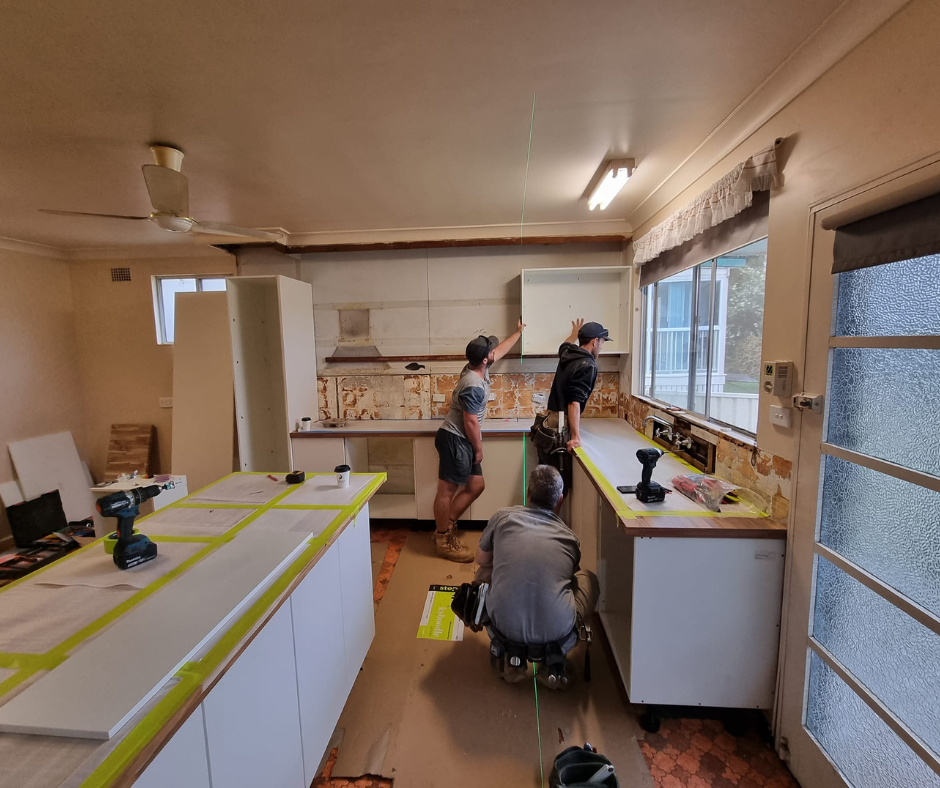 People remodeling a bright kitchen, with cabinets, tools, and masking tape on counters and floor. People remodeling a bright kitchen, with cabinets, tools, and masking tape on counters and floor.
