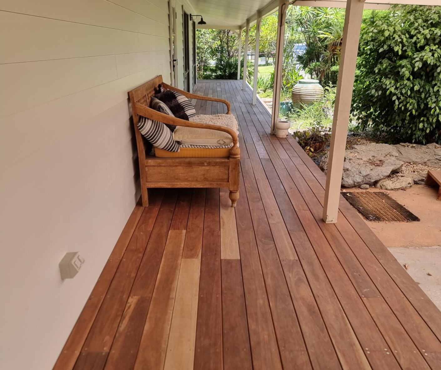 Covered wooden porch with wicker bench, red-toned floorboards, and garden view beyond the railing Covered wooden porch with wicker bench, red-toned floorboards, and garden view beyond the railing