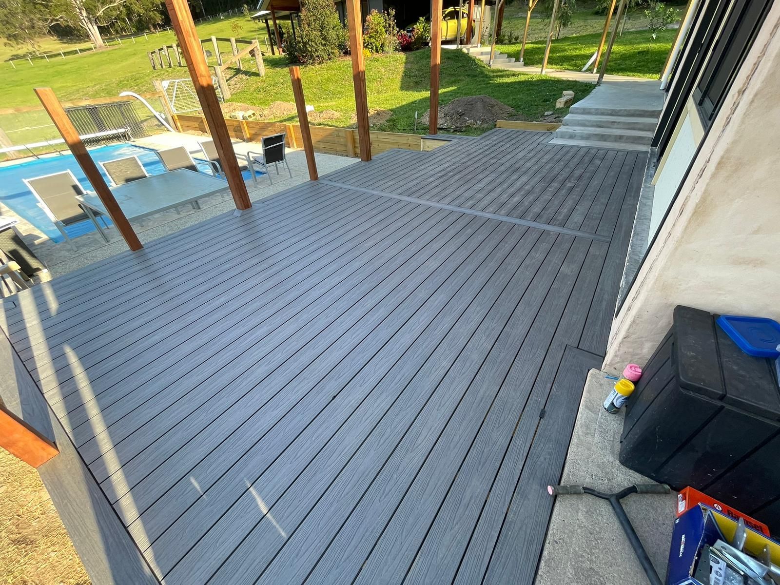 Wooden deck beside a pool and garden path, with lounge chairs and posts under a covered patio.