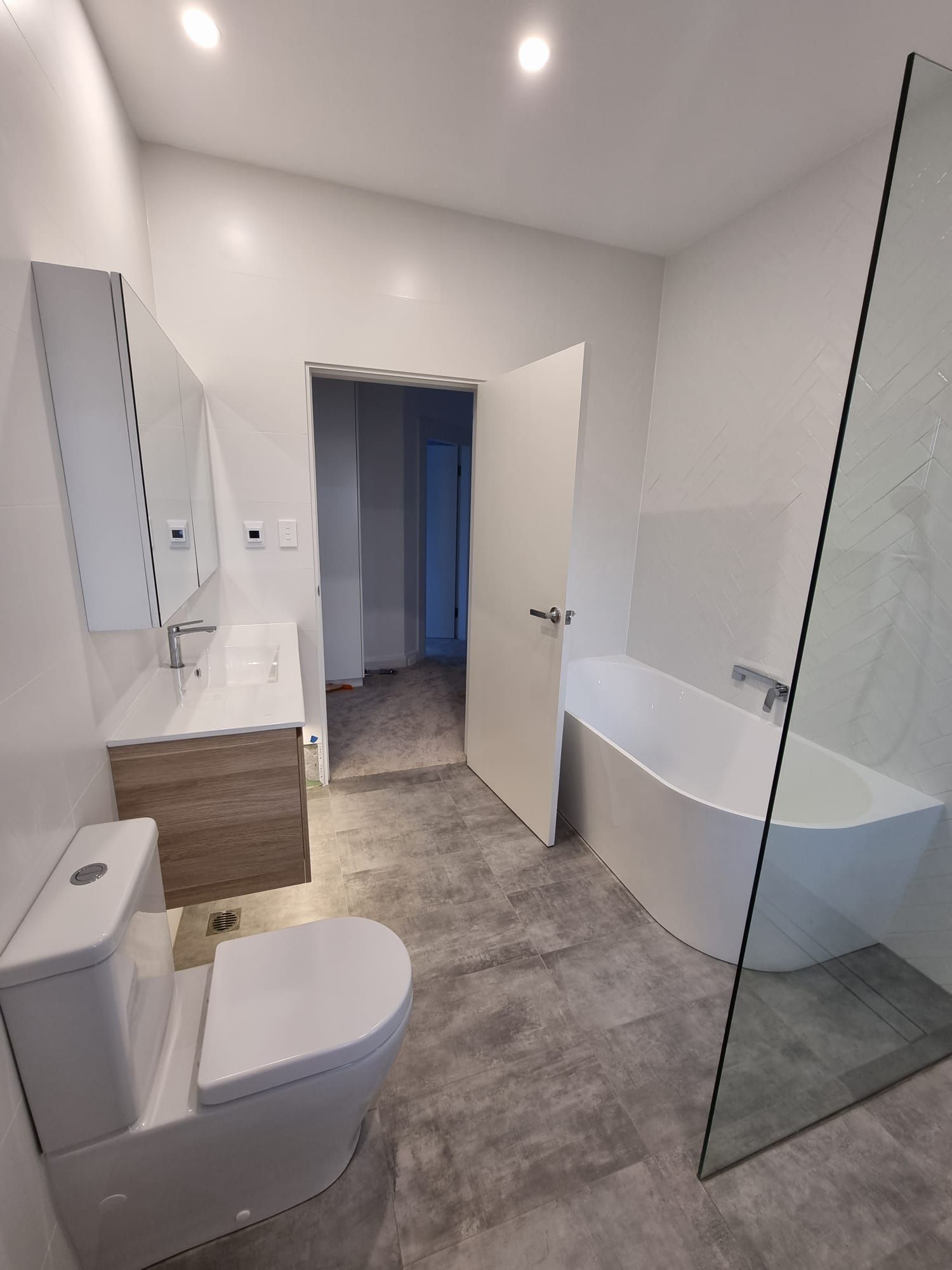 Modern bathroom with a white tub, toilet, and vanity. Gray tile flooring and a glass shower door.
