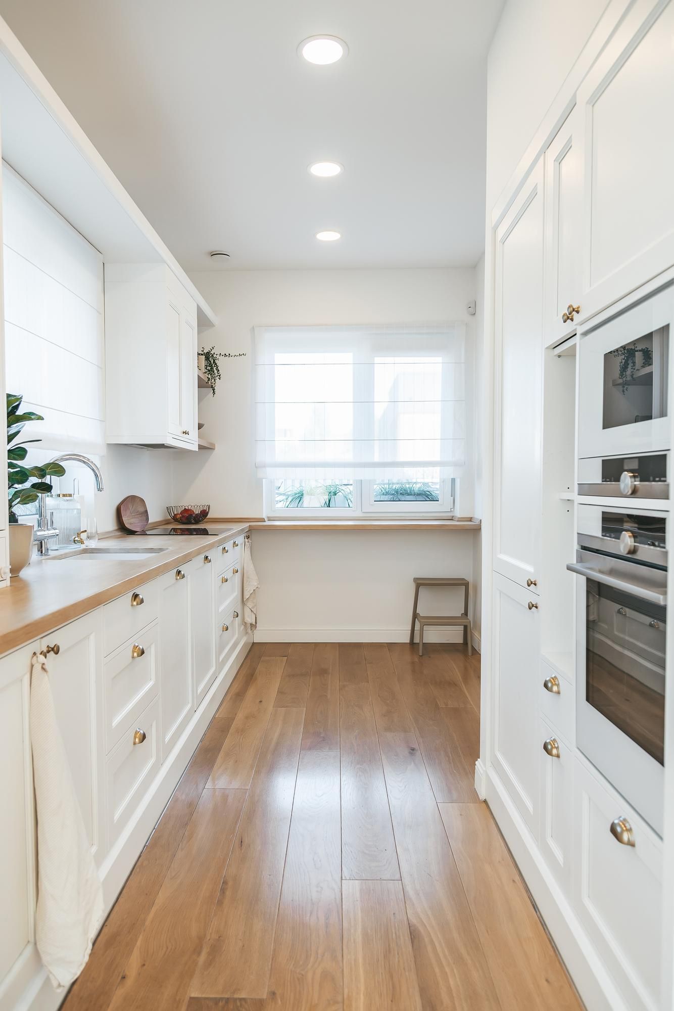 White galley kitchen with wood floor and countertops. Stainless sink, oven, and window.