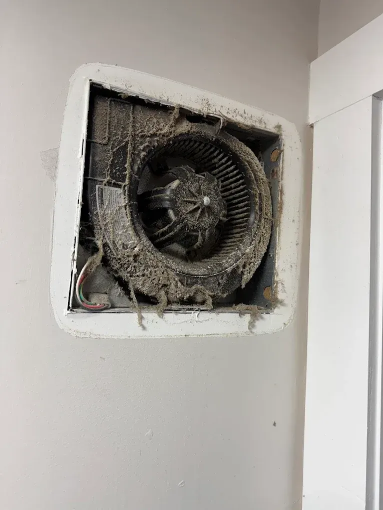Dust-covered bathroom exhaust fan in a white-walled room, next to a white door frame.