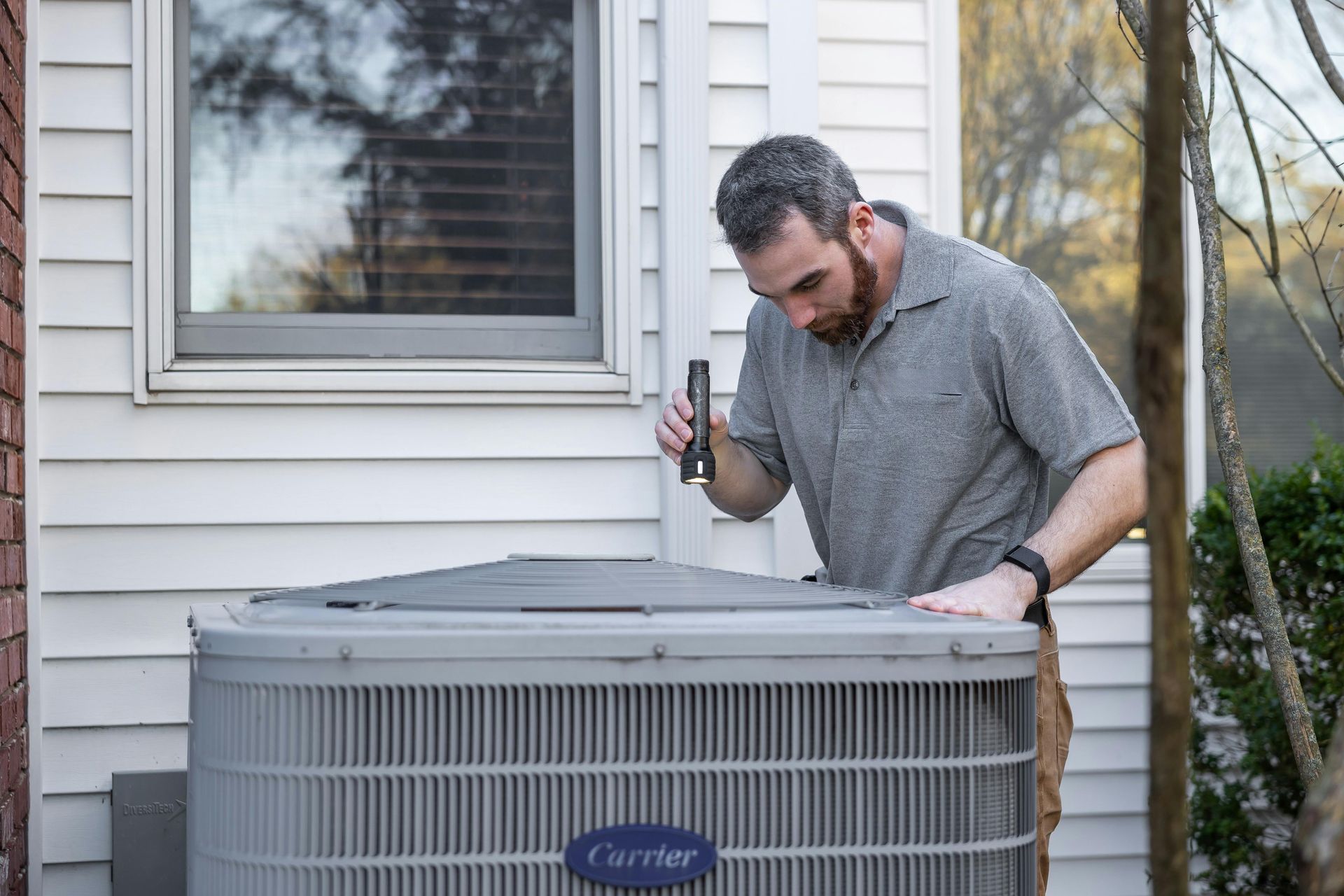 Man inspecting an AC unit with a flashlight outside a house.