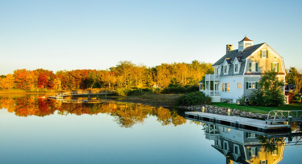 House on a calm lake with a dock, surrounded by trees in autumn colors.