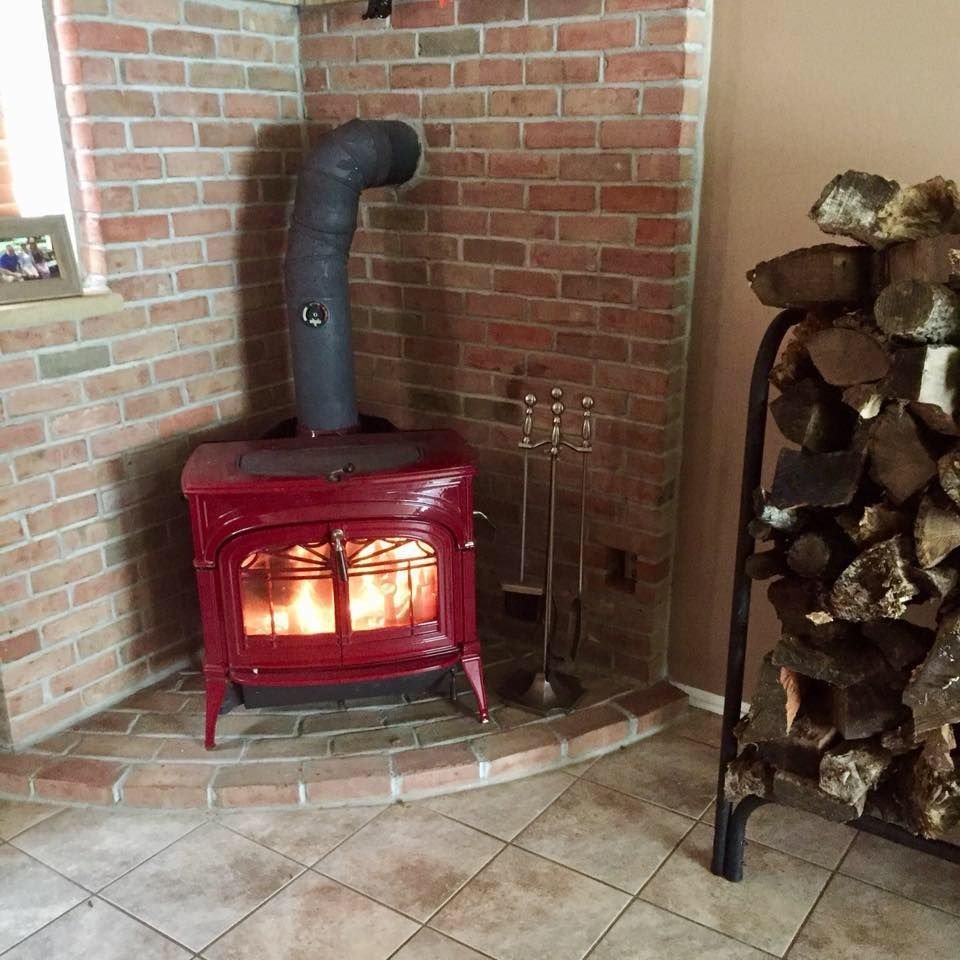 A red wood stove is sitting in the corner of a brick fireplace next to a pile of logs.