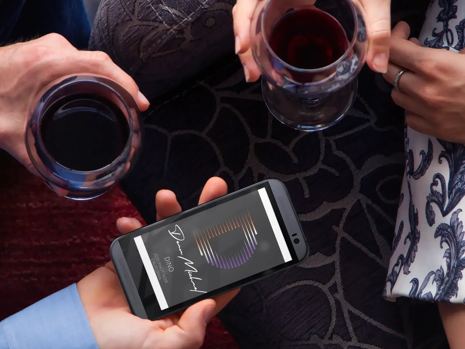 Hands holding wine glasses and a smartphone displaying a logo; dark setting.