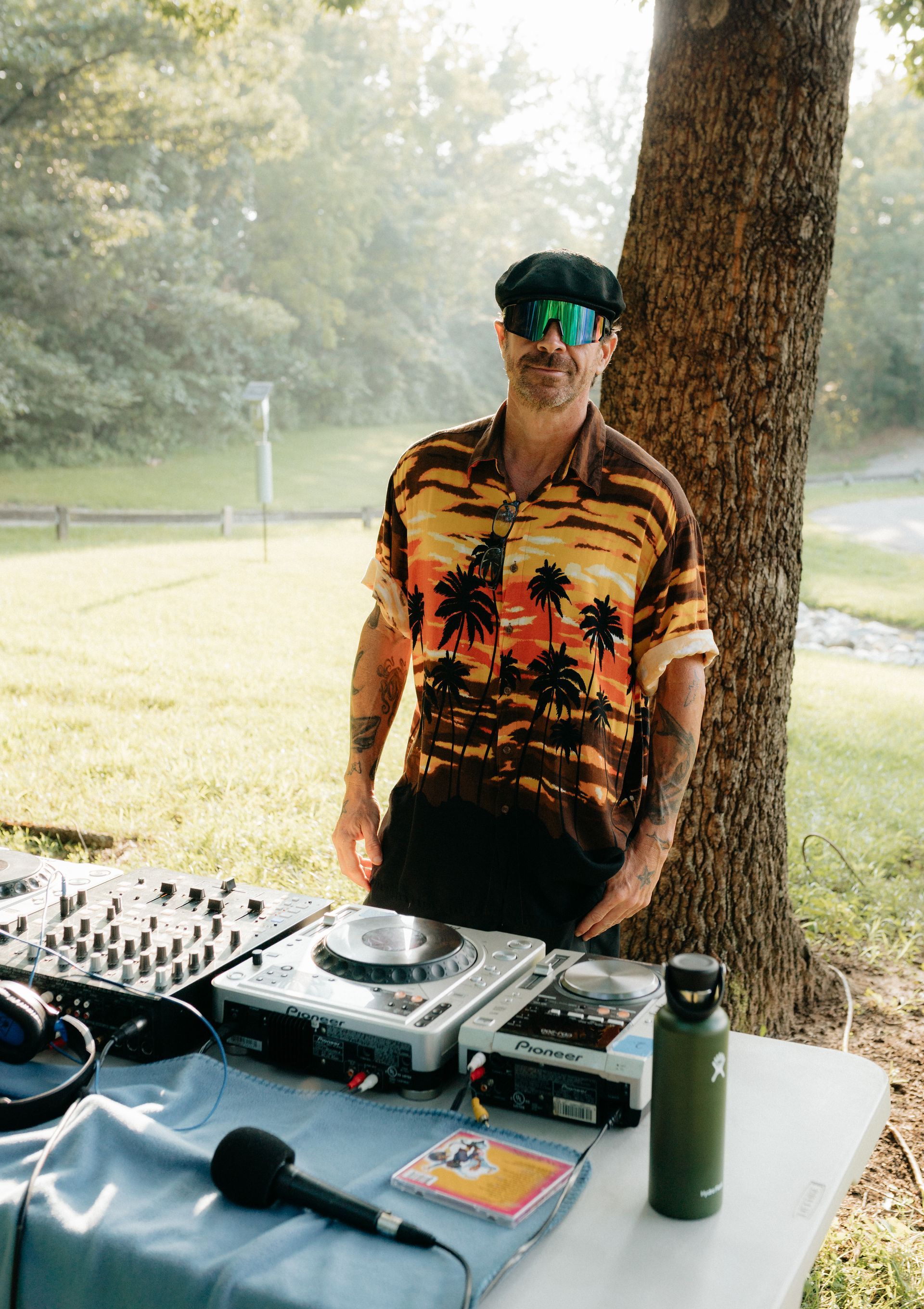 DJ stands outside, set up on a table near a tree. Wearing sunglasses and tropical shirt.