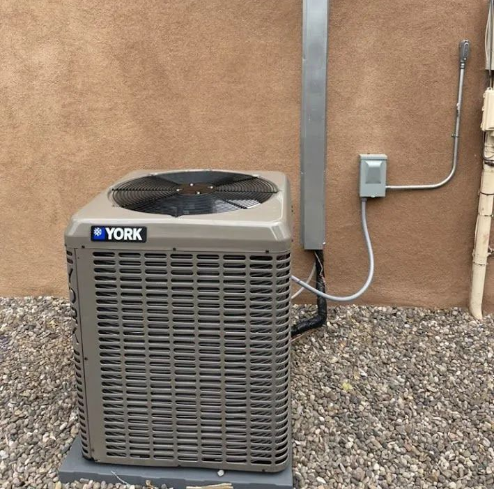 York outdoor air conditioner unit beside a stucco wall and gravel, with exposed conduit and pipes
