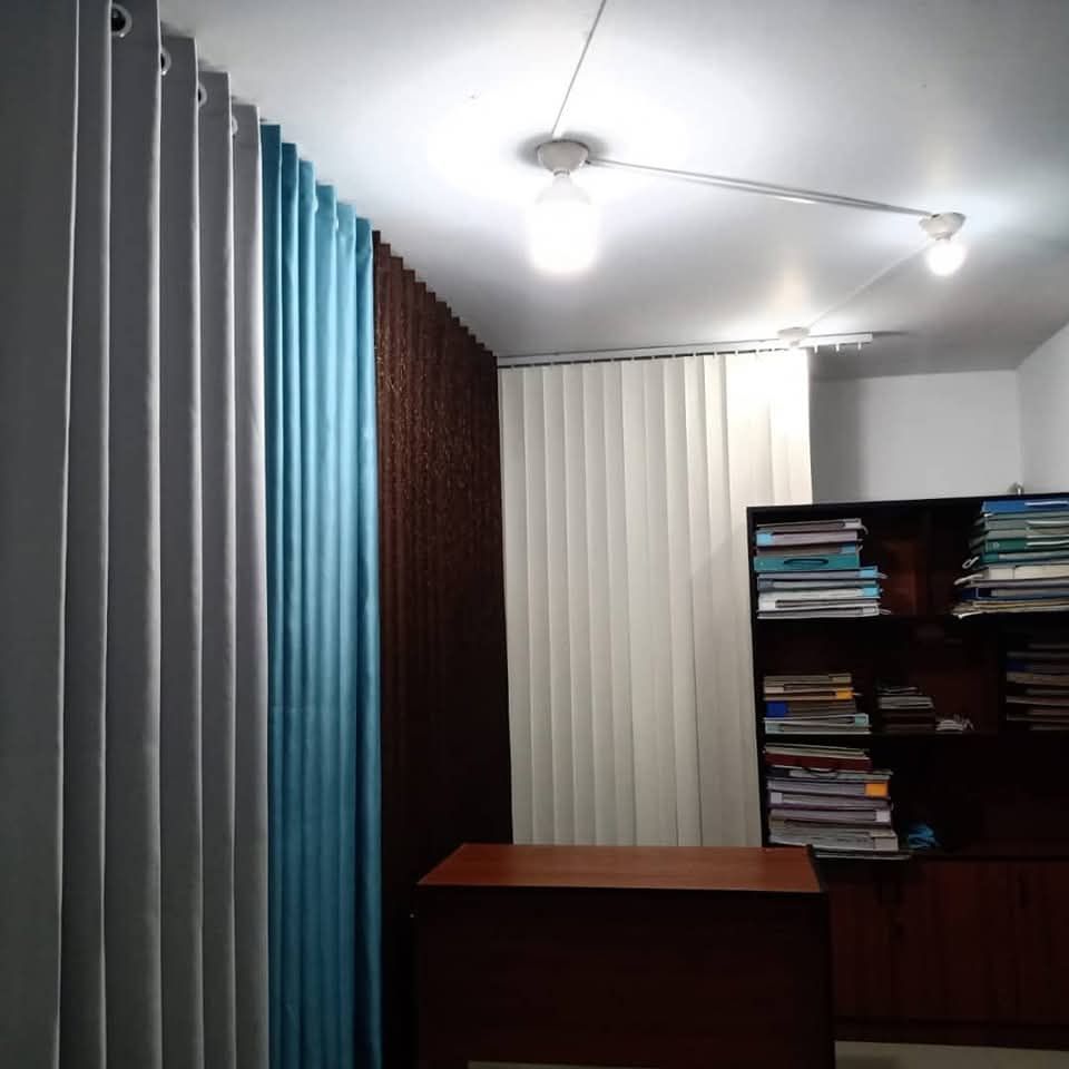 Pequeña oficina con cortinas, un escritorio de madera y una estantería repleta de papeles y libros.