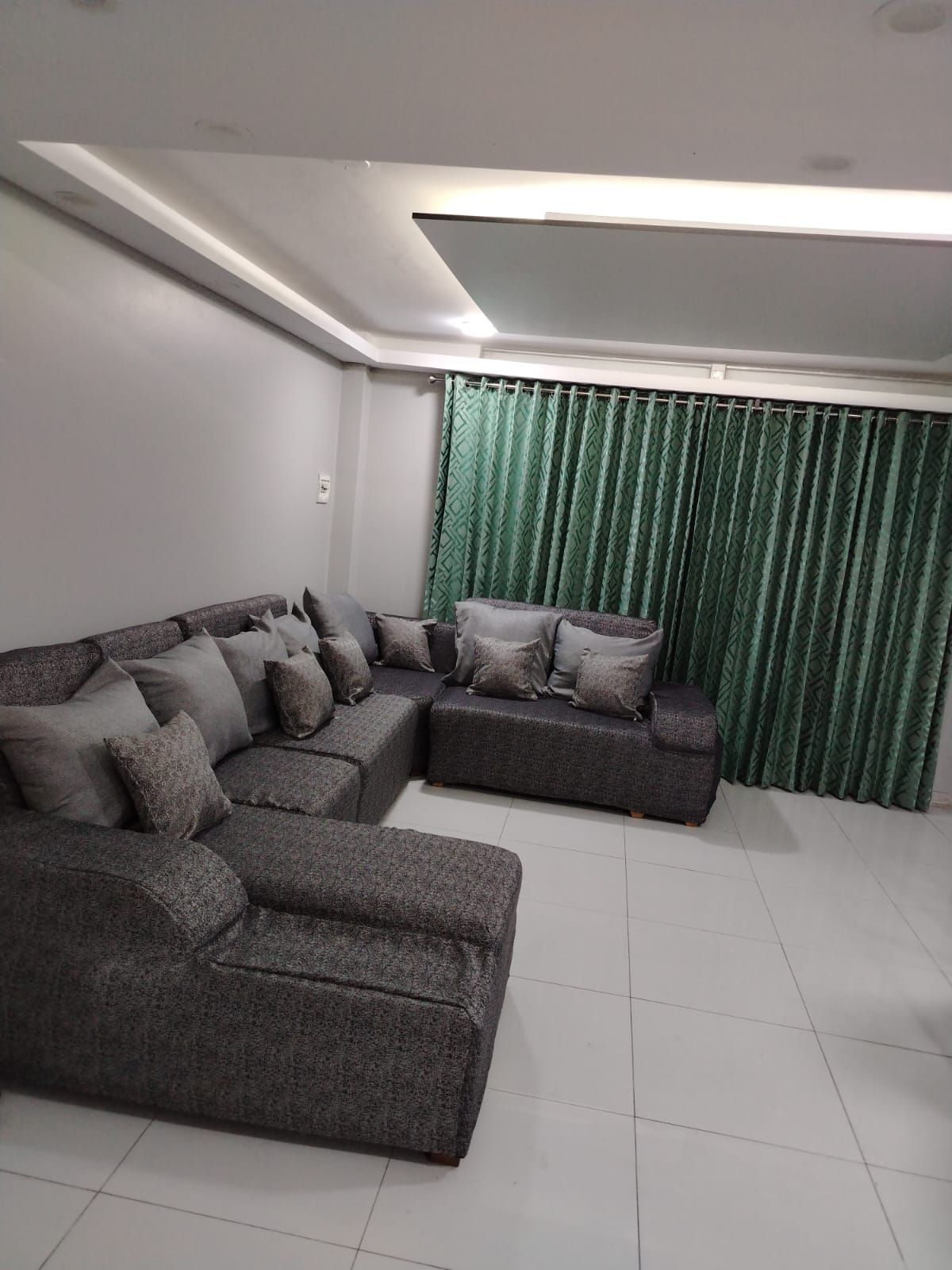 Sala de estar con sofás seccionales grises, suelo de baldosas blancas y cortinas verdes en una ventana.