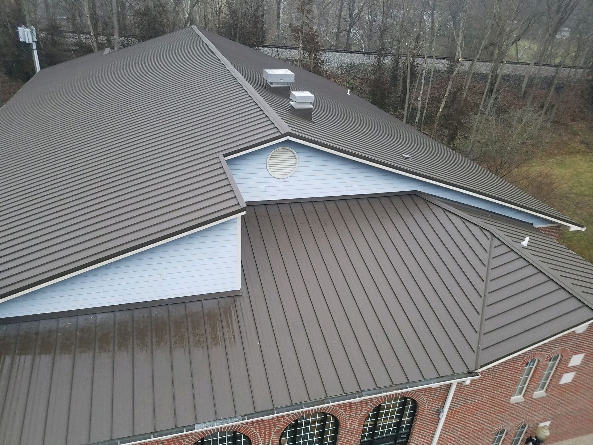 Aerial view of a brick building with a brown metal roof and dormers, surrounded by leafless trees.