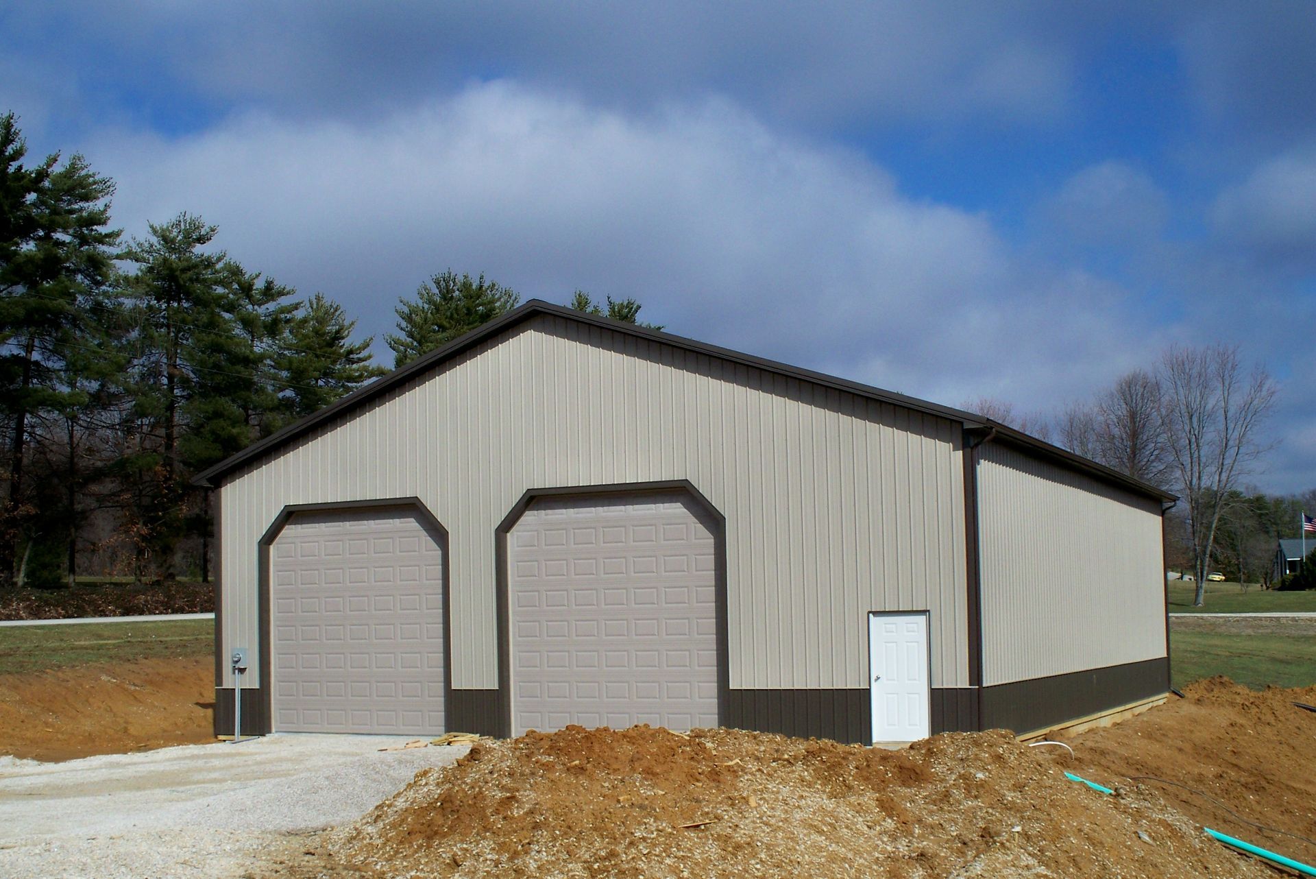 Large beige metal garage with two roll-up doors on a dirt lot under a partly cloudy sky