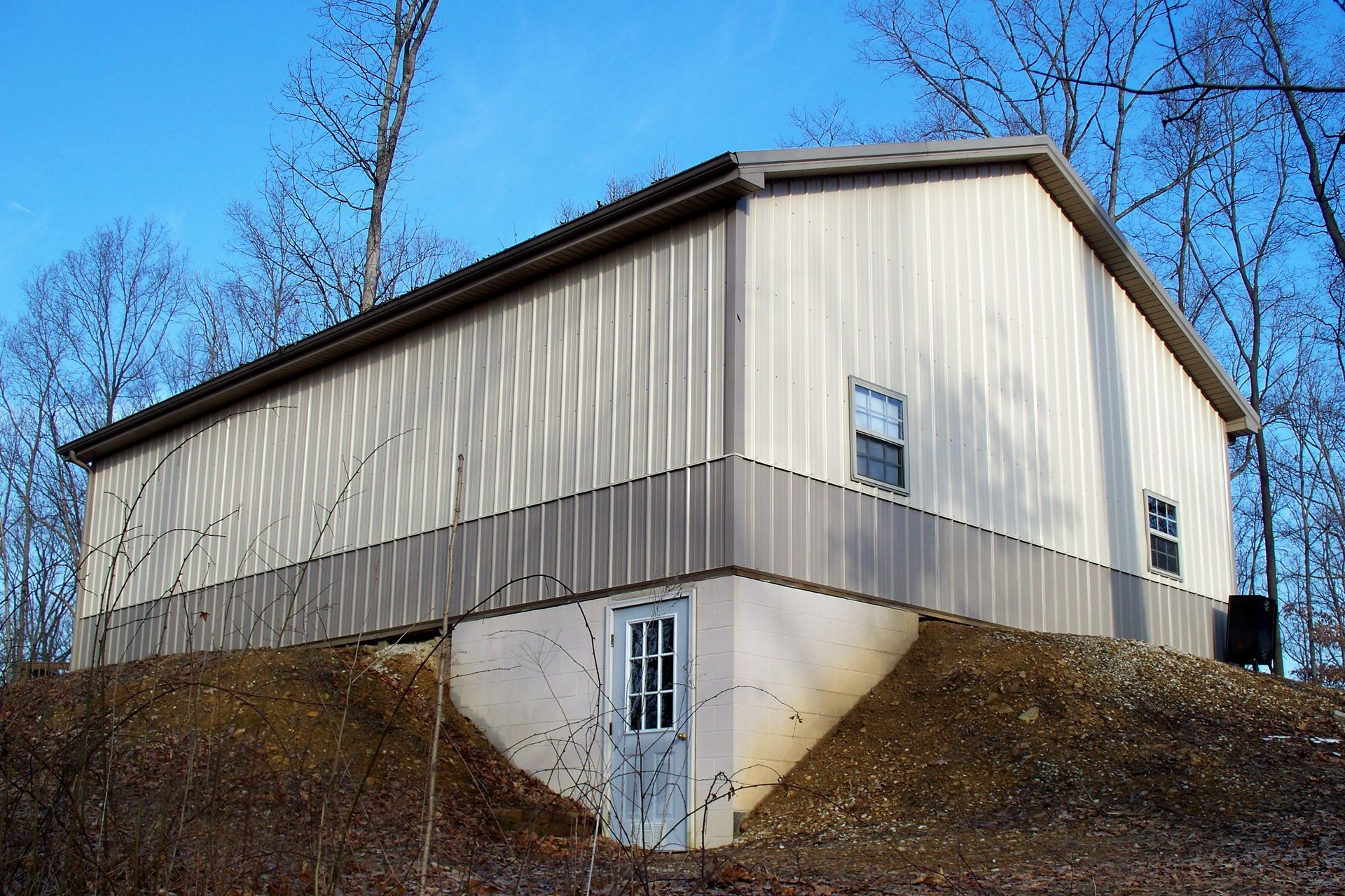 Tan-sided barn on a hillside with a white door and bare trees under a blue sky