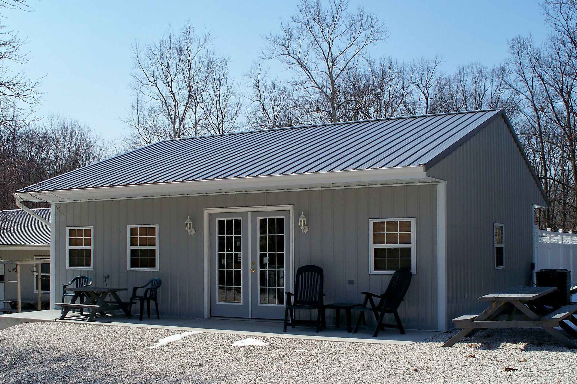 Small gray metal cabin with patio furniture and double glass doors on a gravel lot under bare trees