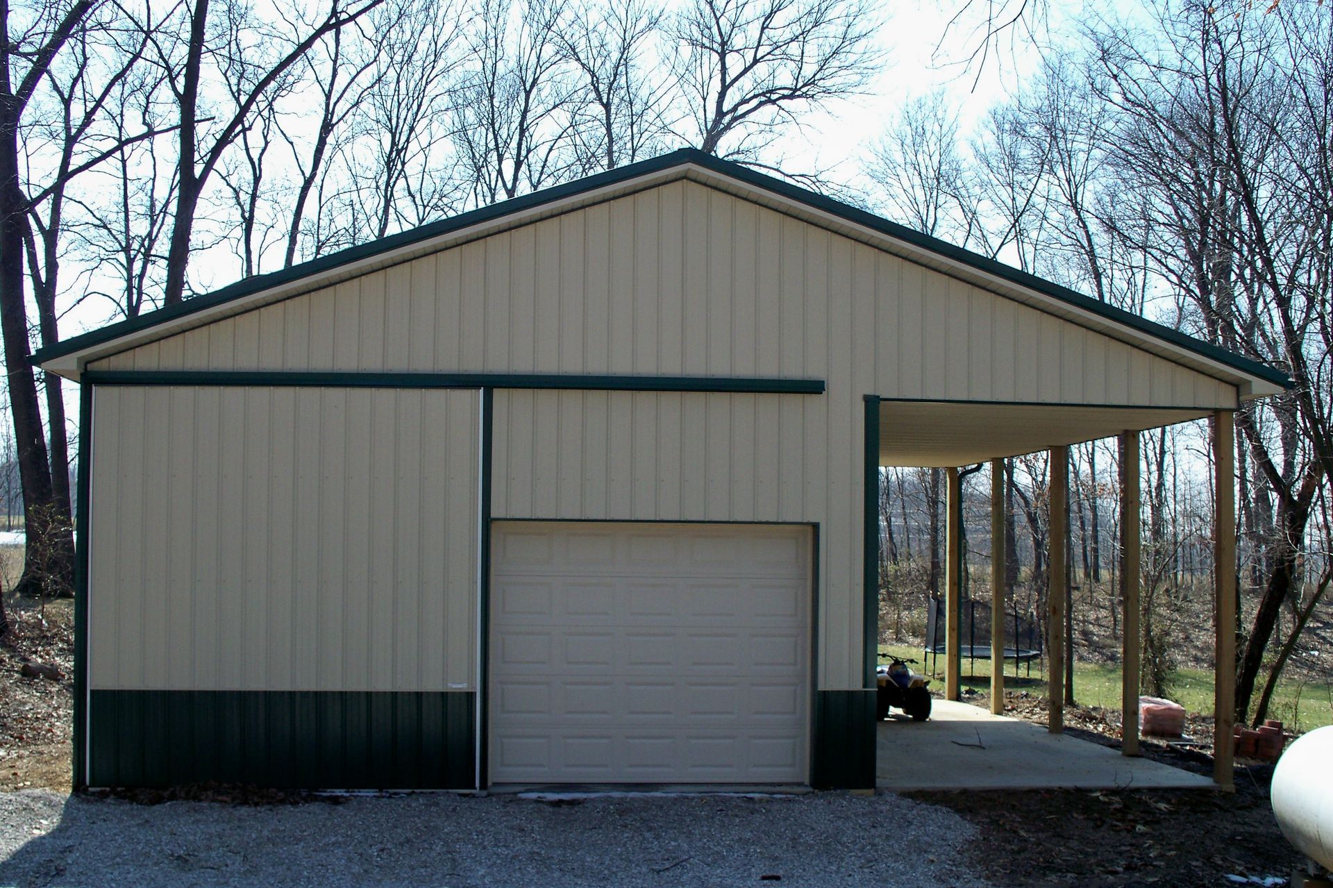 Tan metal shed with a garage door and covered porch in a wooded yard